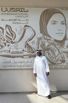 Artista vestido de negro e com barba comprida ao lado de um homem com uma jalaba vestida em frente a um painel em baixo-relevo com motivos alusivos ao mundo árabe e ao automobilismo